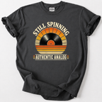 A dark charcoal gray t-shirt with a vintage-style graphic of a vinyl record in a sunset gradient, with the text 'STILL SPINNING' above and 'AUTHENTIC ANALOG' below, laid flat on a light gray background.