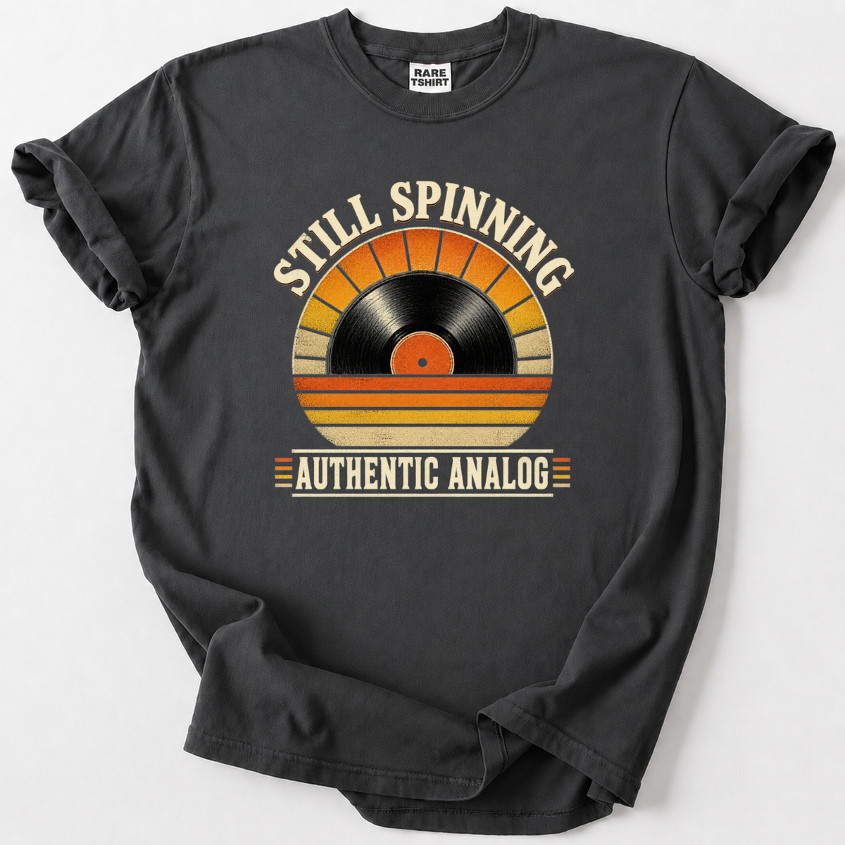 A dark charcoal gray t-shirt with a vintage-style graphic of a vinyl record in a sunset gradient, with the text 'STILL SPINNING' above and 'AUTHENTIC ANALOG' below, laid flat on a light gray background.