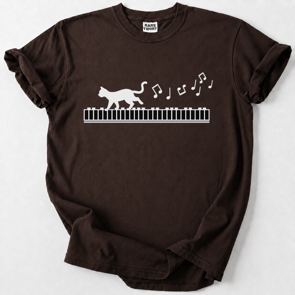A dark brown short-sleeve t-shirt with a white graphic of a cat walking on a piano keyboard and floating musical notes, displayed flat against a light gray background.