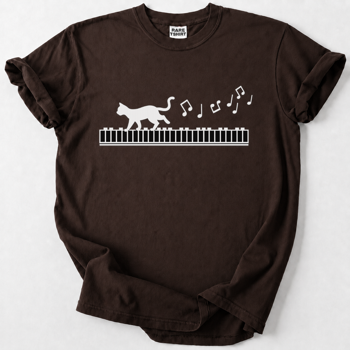 A dark brown short-sleeve t-shirt with a white graphic of a cat walking on a piano keyboard and floating musical notes, displayed flat against a light gray background.